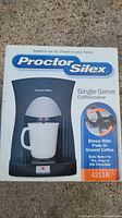Photo of retail packaging showing Proctor Silex Single Serve Coffeemaker model 42114 with features and branding.