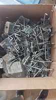 Box filled with numerous silver metal pegboard hooks with double rods and locking mechanism wires.
