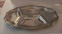 Oval stainless steel serving plate with scalloped edges and three compartments, displayed on table.