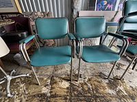 Front view of pair of teal vinyl occasional arm chairs with tubular chrome frames showing both chairs side by side.