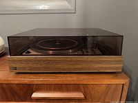 Front view of Dual 1228 turntable with tinted acrylic dust cover in place on dark walnut veneer plinth resting on wooden cabinet.