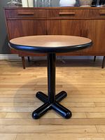 Front view of postmodern round bistro table showing wood tabletop with black border, steel central column and X-shaped steel base, standing on hardwood floor.