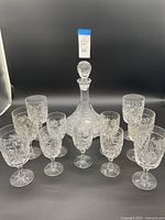 Front view of the crystal decanter with stopper surrounded by ten matching crystal cups in two sizes with cut-glass designs.