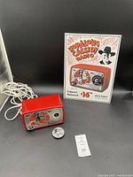 Red vintage Hopalong Cassidy radio with attached power cord, separate tuning knob, and advertising sign in background.