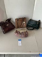 Four ladies handbags arranged on a white table, varying in color and styles: dark brown faux crocodile, medium brown textured, green leather, and small multicolored clutch with bow.