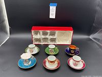 Full view of 6 demitasse cup and saucer sets placed in front of original red presentation box on black background.