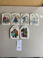 Six decorative wall tiles laid flat on a table, showing colorful folk art characters and nature motifs, each tile has a hole at the top with string for hanging.