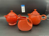 Photo showing three orange ceramic Fiestaware pieces: a lidded sugar bowl, a water pitcher, and a teapot, all with bright orange glossy enamel finish.