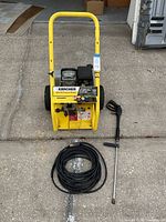 Full view of yellow Karcher pressure washer with pressure hose coiled in front and spray wand beside it on concrete ground.