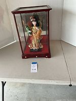 Photo of vintage Japanese geisha doll inside a four-paneled glass and wooden display case on a folded white table.