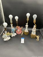 Photo showing four porcelain dresser lamps, including dancing man and woman and two other figures, with light bulbs attached but no shades.