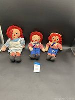 Group photo of three vintage Raggedy Ann and Andy dolls, one larger Ann and two smaller Andy dolls with red yarn hair and classic clothing