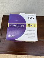 Boxed Sunny Health & Fitness exercise ball, 65 cm, purple color, on wooden table against white wall.