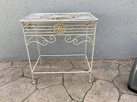 Front view of the ornate metal table showing decorative round floral motif and scrollwork below the slatted metal top.