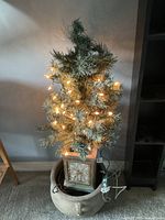 Photo of small artificial pine tree with frosted tips, lit with warm string lights, mounted on a square decorative porcelain base with floral embossed design.