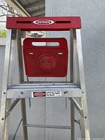 Close-up photo of the top section of the Werner aluminum step ladder showing the red plastic tray and handle, plus warning label on the aluminum spreader.