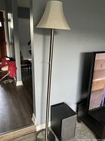 Tall floor lamp standing next to a wall with a beige off-white fabric shade and metal round base showing overall shape and height.