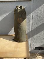 Green camping tent stored upright in a cylindrical green fabric carrying bag, showing minor discoloration due to use or age.