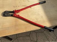 Red bolt cutter hand tool laying on a wooden table with metal cutting head and black rubber handles under natural sunlight.
