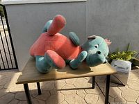 Large plush dinosaur toy laying on its side on a wooden table outdoors, showing coral belly, teal body, and facial features.