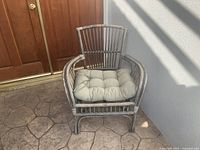 Wicker chair with cushion angled front view showing chair back, arms, cushion, and legs on stone floor with wooden door background.