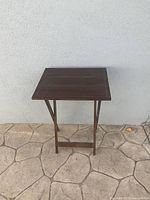 Front view of wooden side table against a light wall, standing on stone pavement, showing square top and leg structure.