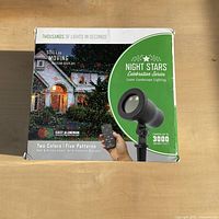 Box front showing Night Stars Celebration Series projector with house illuminated in red and green star patterns, text highlights two colors, five patterns, covers 3000 square feet.