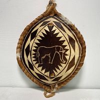 Front view of oval African hide shield with elephant design in center, geometric pattern background and braided edge.