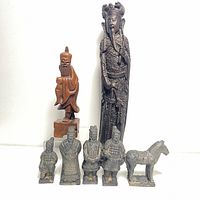 Front view of the five small terracotta warrior figurines with horse, one wooden hand-carved Asian statue, and one large dark resin statue side by side on white background.