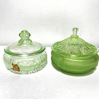 Photo showing two green uranium glass vanity lidded dishes side by side on a white background, highlighting the lid designs and glass textures.