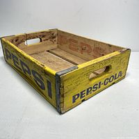 Angled side view showing yellow painted wood with blue Pepsi and Pepsi-Cola branding, metal corner reinforcements, and handle cutouts.