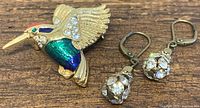 Close-up image of Attwood and Sawyer enamel flying hummingbird brooch with colorful enamel and rhinestones, alongside vintage gold tone rhinestone drop earrings.