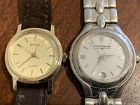 Top-down view showing both watches side by side on a wooden surface, displaying the Birks watch with a leather strap and the Wittnauer watch with a metal bracelet.