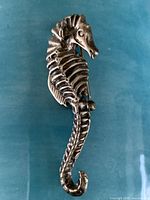 Front view of silver seahorse pin showing detailed body texture and curled tail on a blue background.