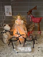 Photo showing group of Christmas decor items on countertop: plush teddy bear with black and white scarf, light-up angel tree topper with lace dress and gold halo spikes, two black metal reindeer candle holders with clear glass cups, red metal reindeer figurine with scarf, and a small white decorative item lying flat.