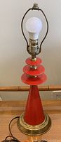 Full view of the lamp showing tomato red painted metal cone base with three circular discs stacked above and brass accents, top socket and harp holder with bulb installed.