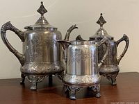 Full set photo showing the tea/coffee pot, creamer, and sugar bowl with ornate designs and footed bases on wooden surface.