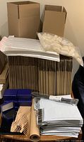 Photo showing large quantity of cardboard boxes, poly bubble mailers stacked around them, roll of brown packing paper, and bag of biodegradable packing peanuts on exposed surface.