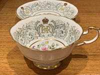 Close-up of the cup showing the gold royal cypher "E II R", crown emblem, blue and grey scrollwork, and central floral design interior.