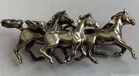 Front view of the detailed silver pin showing three galloping horses in profile, polished sterling silver with fine sculptural detail.