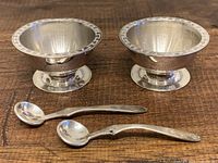 Pair of vintage sterling silver salt and pepper bowls with Greek key decorative rims alongside two matching small sterling silver spoons on wood surface.