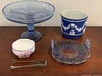 Front view of entire lot showing blue glass cake stand, indigo Wedgwood-style planter, rose sugar bowl, silver tongs and blue glass serving dish