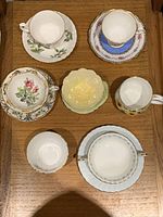 Seven vintage ceramic pieces displayed on wooden surface including cups, saucers, and bowls all with floral and gold detailing.