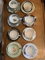 Top-down photo showing eight teacup and saucer sets arranged on wooden surface, showcasing various floral and decorative patterns