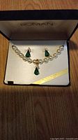 Pearl necklace with green gem and pearl matching earring enhancers displayed inside Roman branded jewelry box on wooden surface.