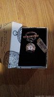 Photo of 1928 Timepieces safety pin pendant with watch charm in branded box, showing front with watch face.