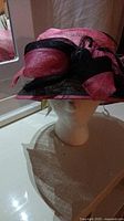 Front view of the pink and black Banash woman's hat on a mannequin head, showing the bow and feather detail.