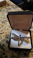 Photo shows dragonfly women's pin in black velvet-lined jewelry box with white interior and gold logo inside lid.