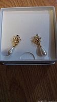 Photo showing brand new Marcella women's earrings in a white jewelry box. Gold-tone with clear rhinestone cluster and teardrop white faux pearls dangling.