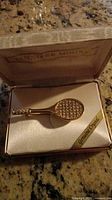 Gold-tone women's tennis racket pin with genuine crystals inside beige presentation box with Jennifer Moore branding.